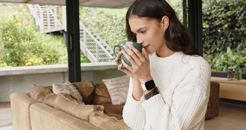Woman enjoying morning coffee while relaxing in cozy sunlit living room with smartwatch