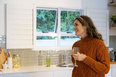 Smiling woman in terracotta cable knit sweater holding glass of white wine in cozy kitchen