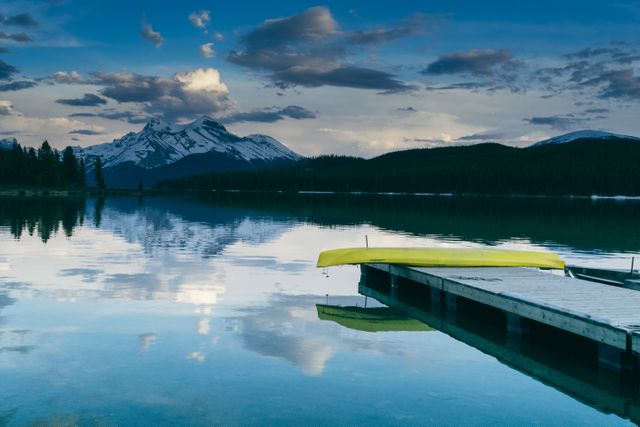 Calm mountain lake at sunset with yellow kayak on dock, reflecting sky and mountains in clear water. Ideal for travel brochures, nature magazines, outdoor adventure ads, or websites promoting ecotourism. Invokes sense of serenity and natural beauty suitable for wallpapers, backgrounds, or promotional materials for leisurely activities.