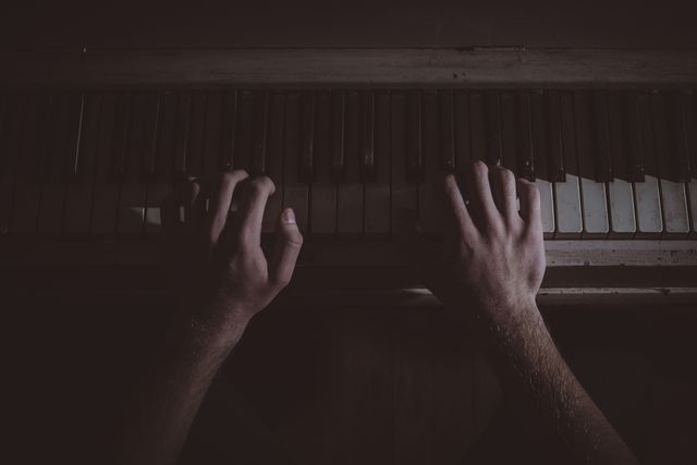 Hands playing on piano keys in a dark, low-light environment, capturing an intimate and creative moment. Perfect for use in music-related content, articles about musicianship, practice, creativity, or vintage-themed projects and promotions.