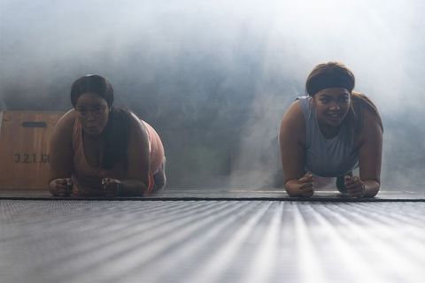 African american women holding forearm plank together on mat in moody backlit gym