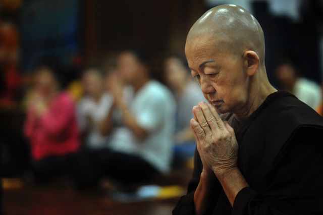 Monk seated, meditating and praying in temple surrounded by people. Ideal for use in stories about spirituality, Buddhism, meditation practices, inner peace, and religious traditions. Can be used in religious websites, meditation apps, and publications about mindfulness.