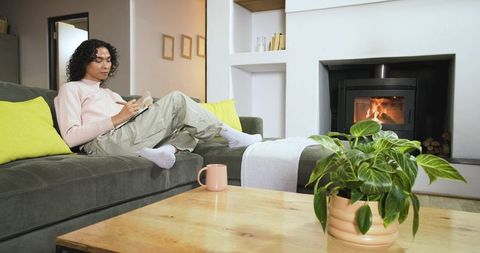 Nonbinary person reclining on sofa writing in notebook by fireplace in cozy living room