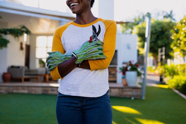 African American woman standing in garden, smiling and holding garden shears while wearing safety gloves. Ideal for content related to gardening, outdoor activities, home improvement, and lifestyle. Perfect for blogs, articles, and advertisements promoting gardening tools, outdoor living, and healthy hobbies.