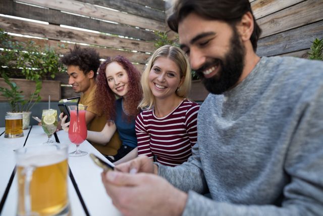 Group of friends enjoying time together at an outdoor bar, using mobile phones while smiling and socializing. Ideal for themes related to friendship, technology, social media, leisure activities, and modern lifestyle.