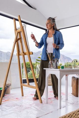 Senior woman artist painting barefoot in bright studio with easel and mountain view