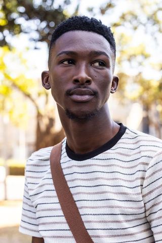 Young african man looking left wearing striped tee and crossbody bag in sunlit park
