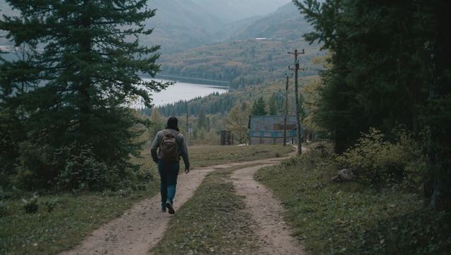 Solo hiker walking along rutted mountain track toward lakeside cabin in autumn valley