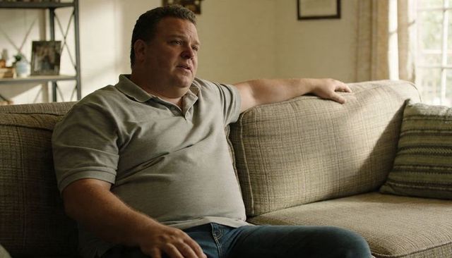 Middle-aged man relaxing on beige sofa in cozy sunlit living room wearing polo and jeans