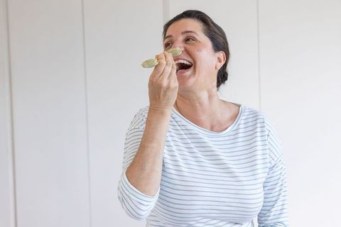 Mature woman laughing while tasting with spoon, lifting utensil toward mouth at home