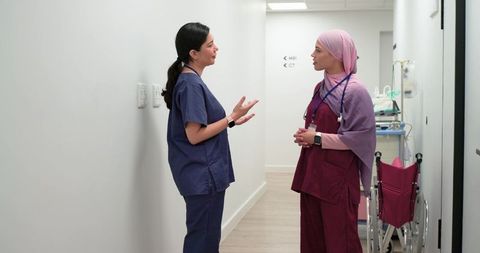 Female nurses in scrubs and hijab conversing in hospital corridor near mri ct and wheelchair