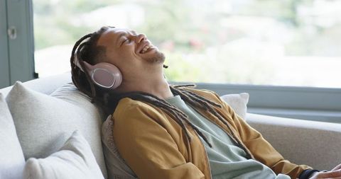Young man with dreadlocks relaxing on sofa listening to music with pink headphones