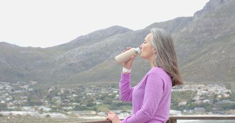 Senior woman drinking from reusable bottle on coastal overlook with mountain town backdrop