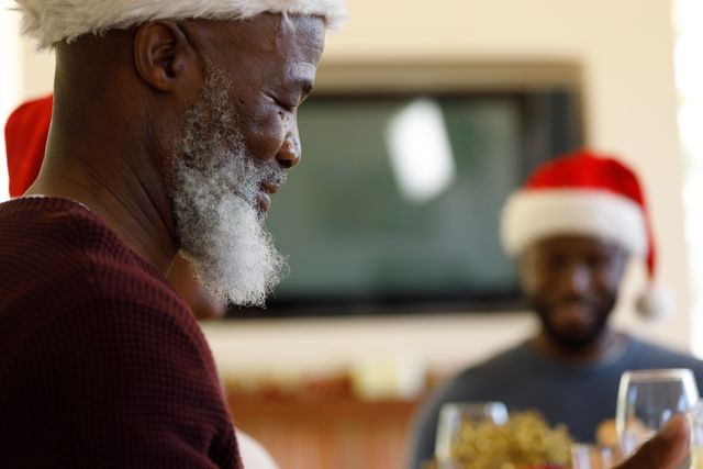 Close up of an old african-american man eating with his family on a christmas dinner table with all of them wearing santa hats. wine glasses can be seen on the table.