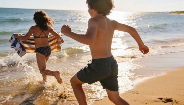 Kids running and splashing on sunlit shore holding striped towel for summer fun