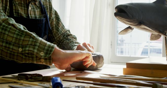 Craftsperson working on wooden fish sculpture with various tools on wooden table. Ideal for use in articles or advertisements focused on creativity, art, craftsmanship, woodworking, and hobbies.
