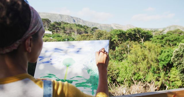 Image depicts an artist painting a landscape outdoors amidst greenery and mountains. The artist is focusing on their canvas, capturing the natural scenery. Ideal for illustrating themes of creativity, art in nature, outdoor activities, artistic hobbies, and the beauty of the outdoors.