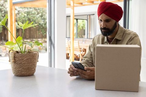 Man in red turban checks smartphone beside parcel in bright kitchen