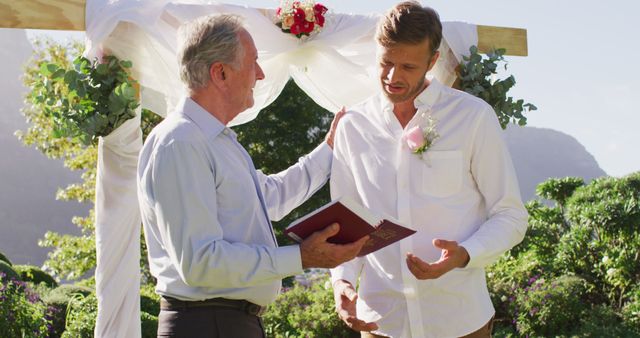 Smiling caucasian senior male wedding officiant holding book and groom standing in outdoor altar. romantic summer wedding outdoors.