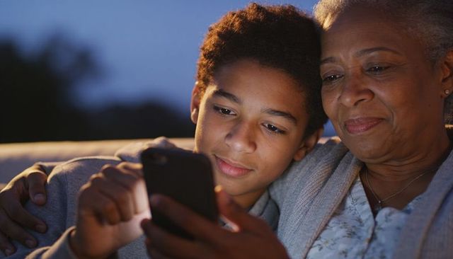 Grandmother and grandson sharing smartphone on couch, evening bonding and warmth