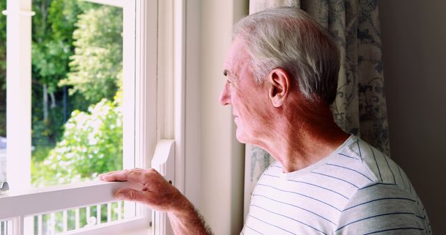Senior man stands by a window, appearing reflective and deep in thought. This captures a moment of quiet contemplation, highlighting themes of loneliness and introspection. Could be used in contexts involving elderly well-being, dealing with loneliness, mental health, retirement, or isolation.