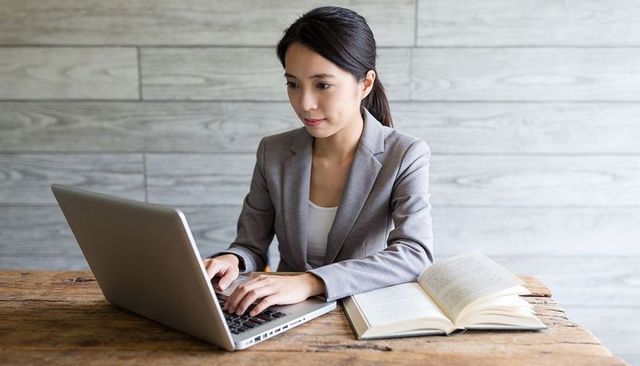 Focused professional woman typing on laptop at rustic wooden desk with open paperback book