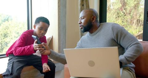 African american father teaching son smartphone and laptop learning in sunlit living room