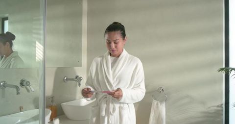 Mid-adult asian woman examining pregnancy test in minimalist bathroom wearing white robe