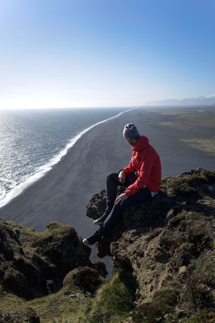 Individual wearing warm clothes sitting on cliff edge, looking at expansive ocean view with stretching black sand beach below. Perfect for articles or promotions on travel, adventure, coastal destinations, nature exploration, or advertising outdoor apparel and gear.
