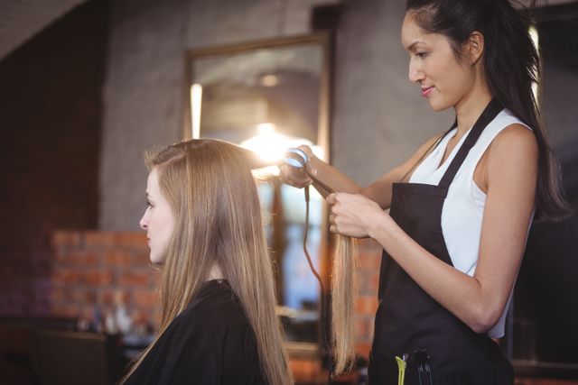 Hairdresser straightening client's hair in a salon. Ideal for use in beauty and hair care advertisements, salon promotions, hairstylist training materials, and professional service websites.