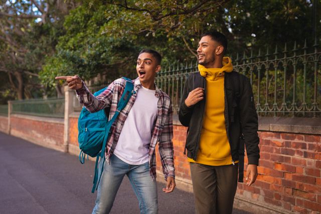 Two happy biracial male friends with backpacks are walking and talking in a city street. They appear to be enjoying their travel break, exploring the urban environment. This image is perfect for promoting travel, tourism, friendship, and adventure. It can be used in advertisements, travel blogs, social media posts, and brochures.