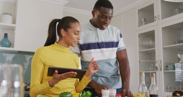 Depicts a cheerful couple engaging in meal preparation in a well-decorated modern kitchen. They are working together, using a tablet for recipes or instructions, surrounded by fresh vegetables and utensils. Ideal for themes related to domestic life, cooking, healthy living, technology in everyday tasks, and family bonding.