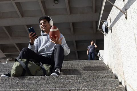 Teen male sitting on stadium steps holding football checking smartphone wearing headphones