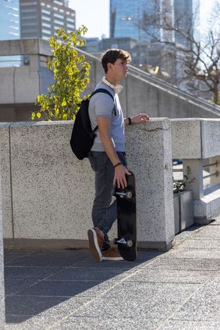 Sunlit urban teen leaning on parapet holding skateboard, backpack, headphones, downtown