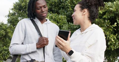 Diverse friends sharing smartphone and chatting while wearing hoodies outdoors