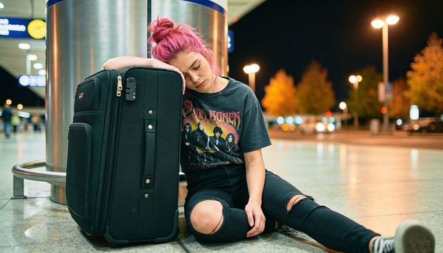 Pink-haired traveler leaning on suitcase at airport curb at night wearing band tee