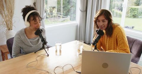 Diverse female co-hosts recording casual home podcast with laptop, microphones, headphones