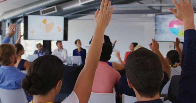 A group of business professionals attend a seminar or conference. Some audience members are raising their hands to ask questions. Presentation involves pie charts displayed on screens. This image is useful for illustrating corporate seminars, training events, company meetings, professional development, and interactive business conferences.