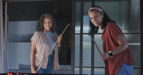 Women playing pool in home game room laughing aiming shot cue sticks casual companions