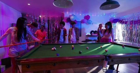 Diverse friends playing pool at neon party with balloons, tinsel and festive lighting