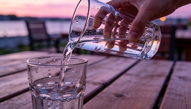 Hand pouring water from glass pitcher into glass on wooden table at sunset by waterfront