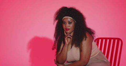Mid-adult african american woman sitting thoughtfully on red chair against pink backdrop