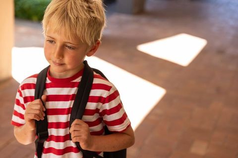 Blonde boy standing with backpack in sunlit arcade wearing red striped shirt