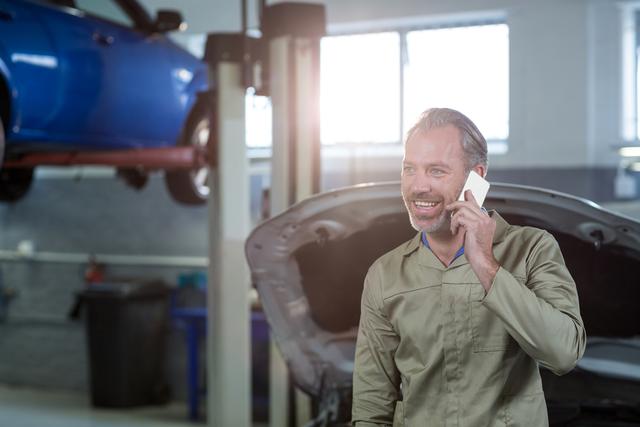 Mechanic smiling while talking on mobile phone in auto repair shop. Ideal for illustrating automotive services, customer interaction, professional mechanics, vehicle maintenance tasks, and repair workshops.
