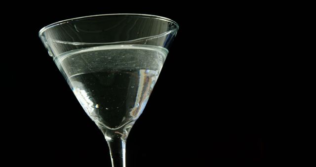 A martini glass is partially filled with a clear liquid, set against a dark background, with copy space. Its elegant shape and the simplicity of the composition suggest a sophisticated atmosphere ideal for a beverage-related theme.