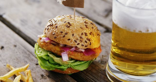 A delicious chicken burger with lettuce and red onions served on a sesame seed bun, alongside a glass of cold beer on a rustic wooden table. There are also some fries off to the side. This setup is perfect for use in food blogs, menus, restaurant advertisements, and social media posts to showcase casual dining and comfort food options.