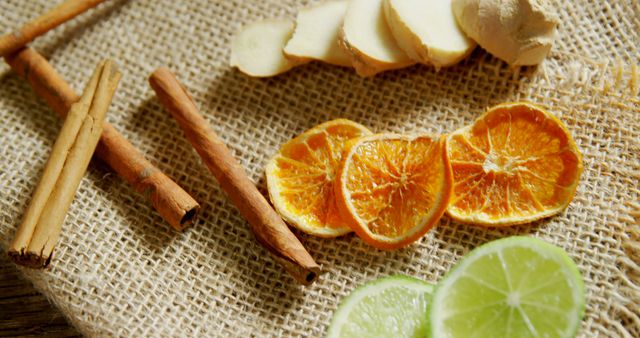 Slices of dried orange, fresh lime, ginger, and cinnamon sticks are arranged on a burlap surface, with copy space. These ingredients are often used in cooking and natural remedies, adding a burst of flavor and aroma.