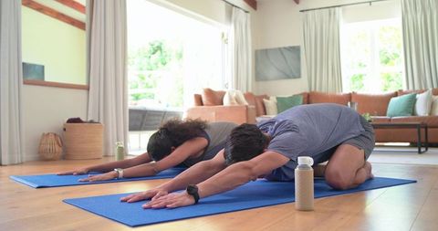 Couple practicing yoga stretch on blue mats in sunlit living room with water bottles