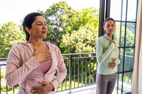 Two women practicing mindful breathing on sunny balcony with lush greenery and natural light