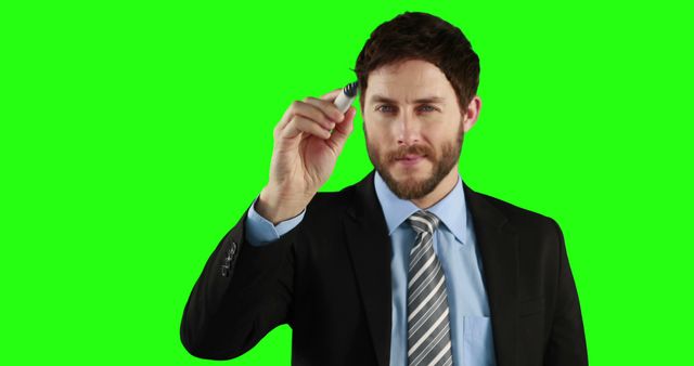 Young businessman in formal suit holding marker and pointing as if writing or drawing, posing against green screen background. Suitable for use in presentations, advertisements, marketing materials, educational videos, or corporate training content.